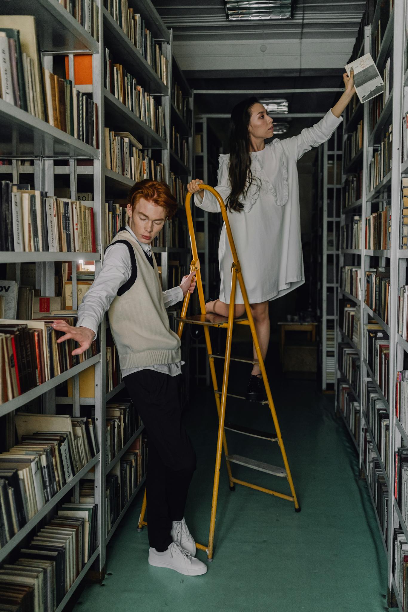 Two young adults in a university library browsing books with a ladder.